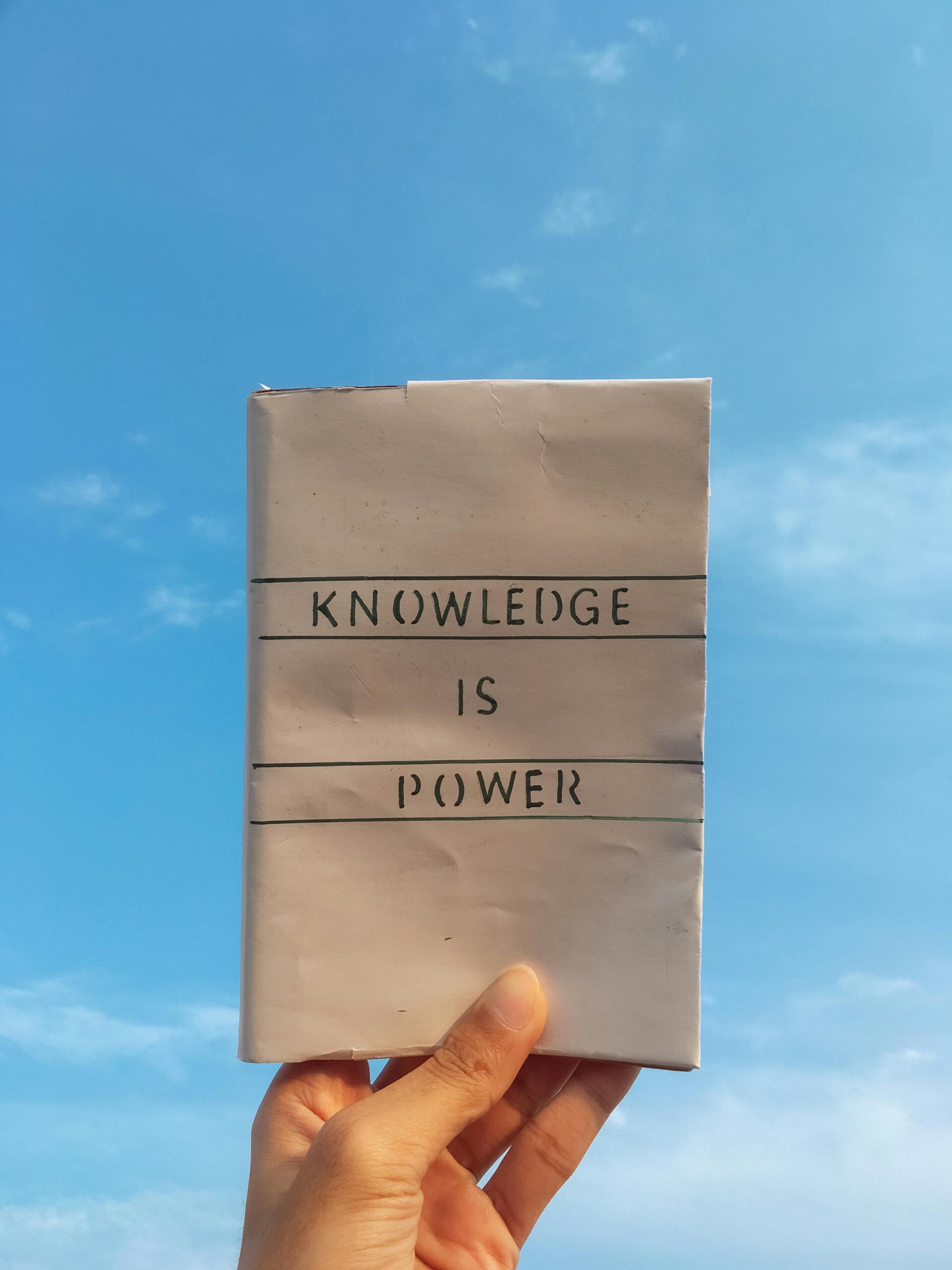 pexels-photo-7949588-7949588 A hand holds a book with the quote 'Knowledge is Power' against a clear blue sky.